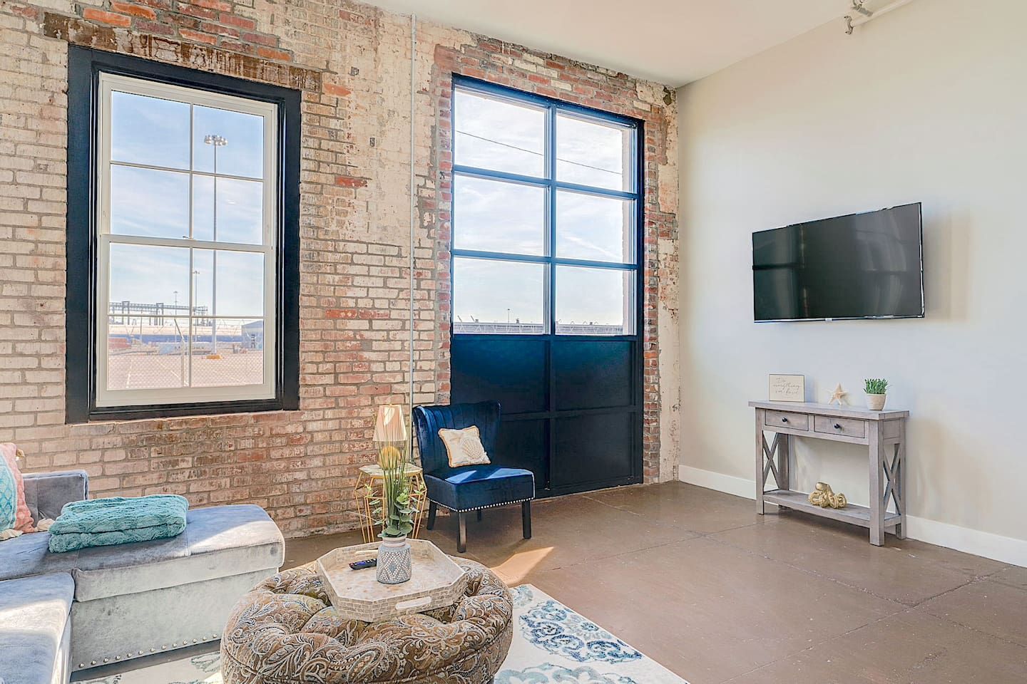 A room with a brick wall, a large window, a blue sofa, a round ottoman, a small table with a plant, and a mounted TV.