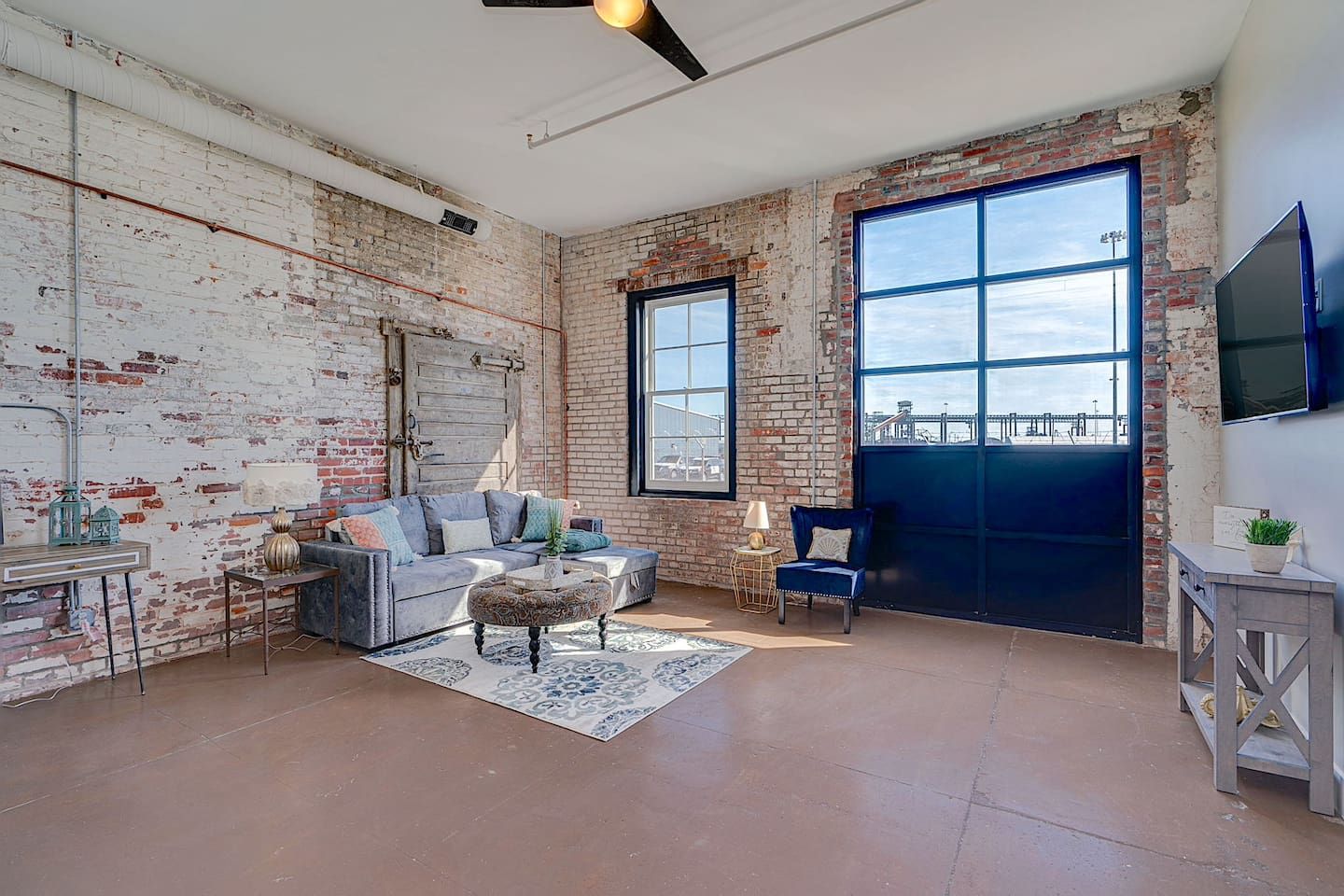 A spacious living room with exposed brick walls, a large window, a sofa, a coffee table, and a television mounted on the wall.