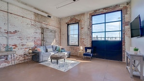 Un salon spacieux avec des murs en briques apparentes, une grande fenêtre, un canapé, une table basse et un téléviseur fixé au mur.