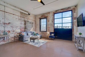 Un salon spacieux avec des murs en briques apparentes, une grande fenêtre et un ventilateur de plafond.