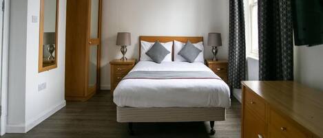 A bedroom with a large bed, two bedside tables with lamps, a wooden dresser, and a mirror on the left wall.