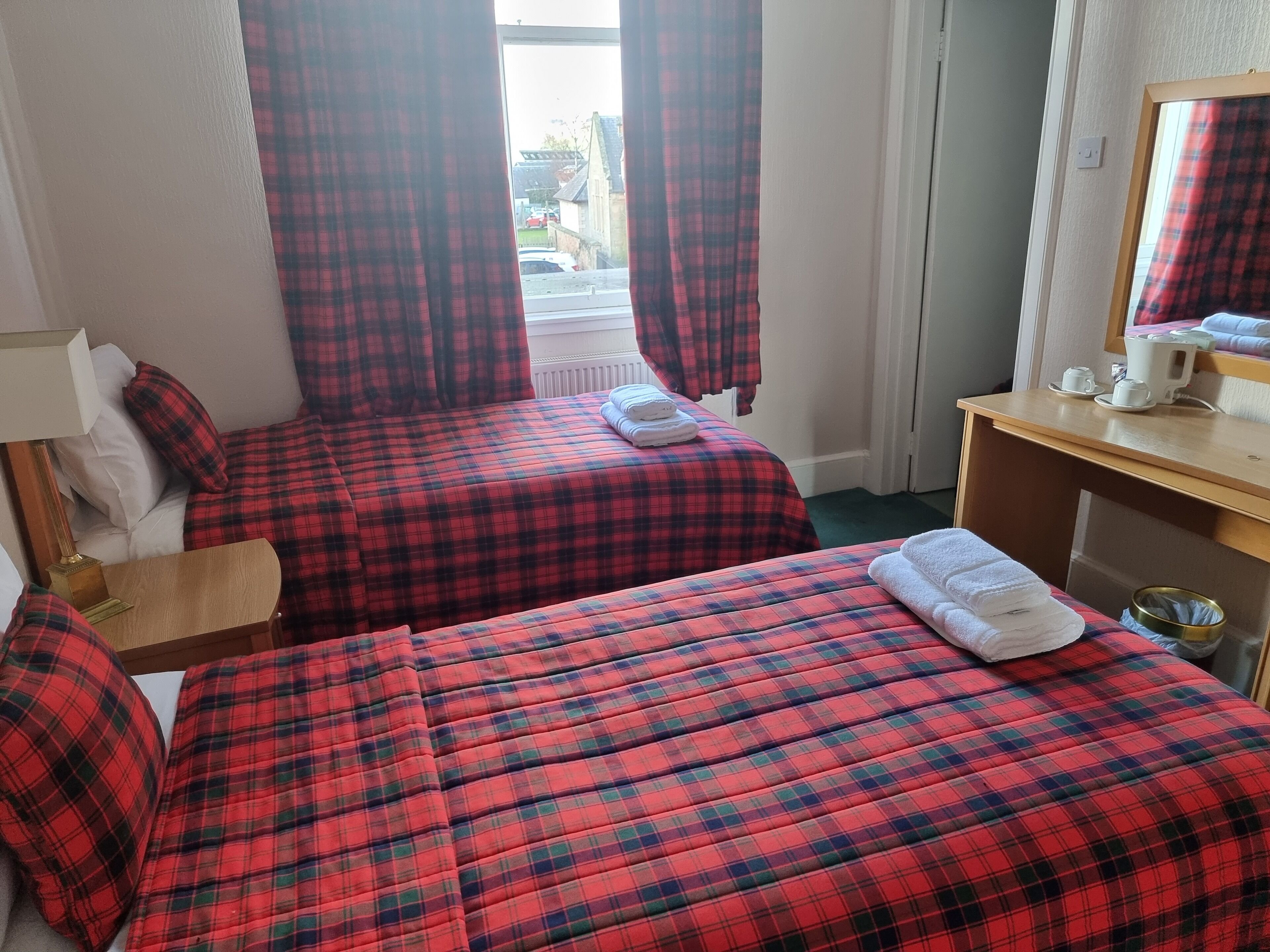 A hotel room with two beds, red and black checkered bedding, a wooden nightstand, a mirror, and a window with curtains.