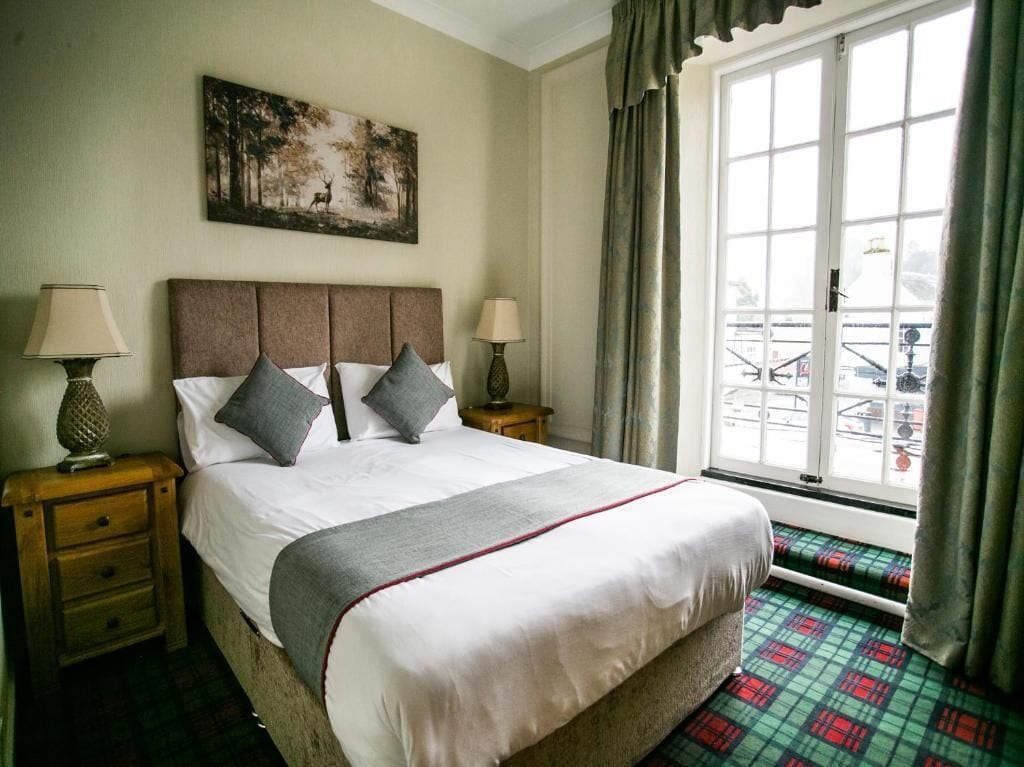 A neatly made bed with pillows, a nightstand with a lamp, a painting on the wall, and a window with curtains.