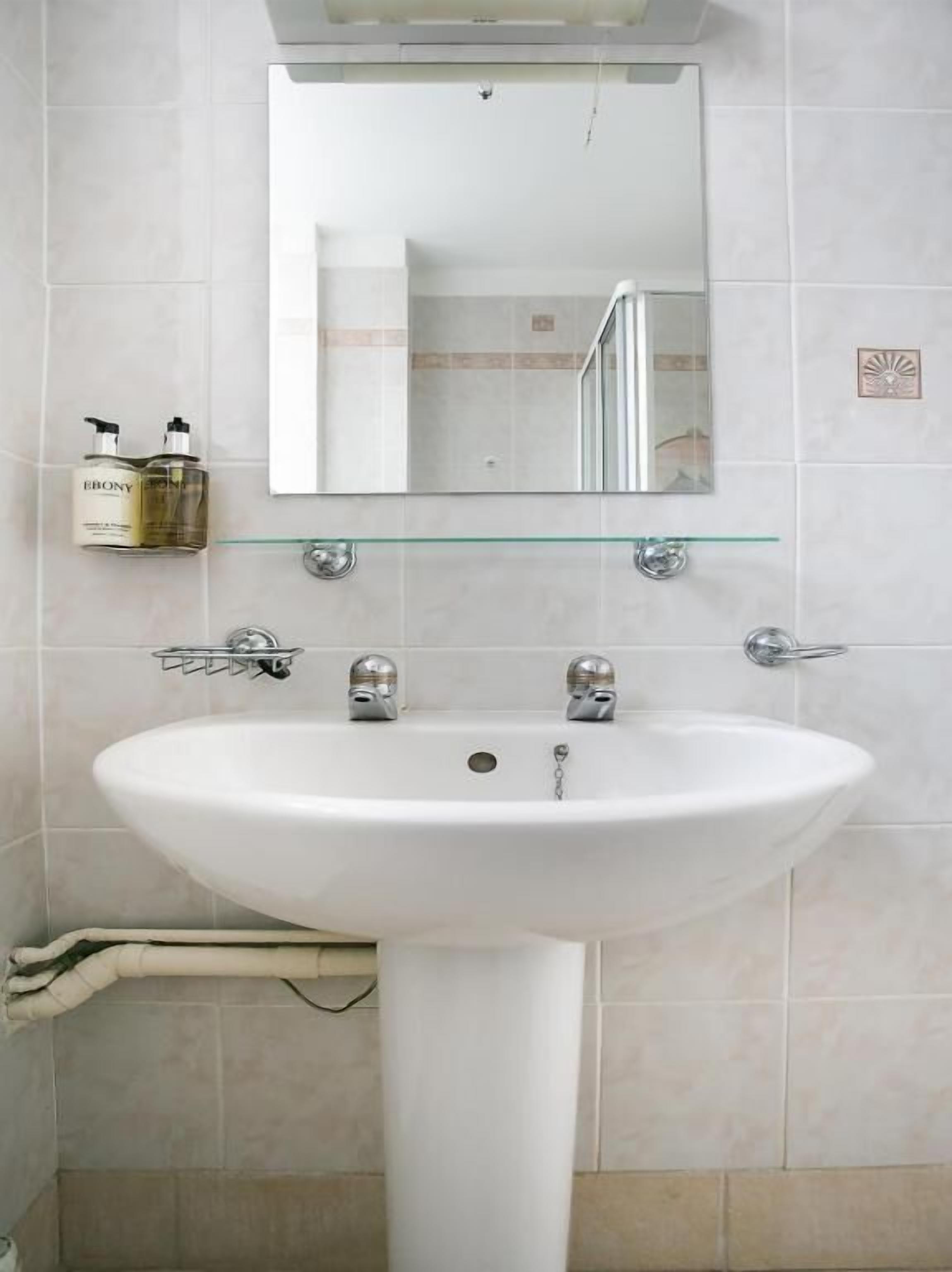 Ein Badezimmer mit einem weißen Waschbecken, zwei Wasserhähnen, einer Glasablage für Toilettenartikel und einem Spiegel über dem Waschbecken.