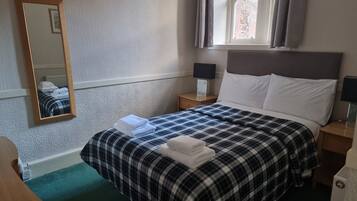 Una cama bien hecha con una colcha de cuadros y almohadas blancas, una mesita de noche con una lámpara, un espejo en la pared y una ventana con cortinas.