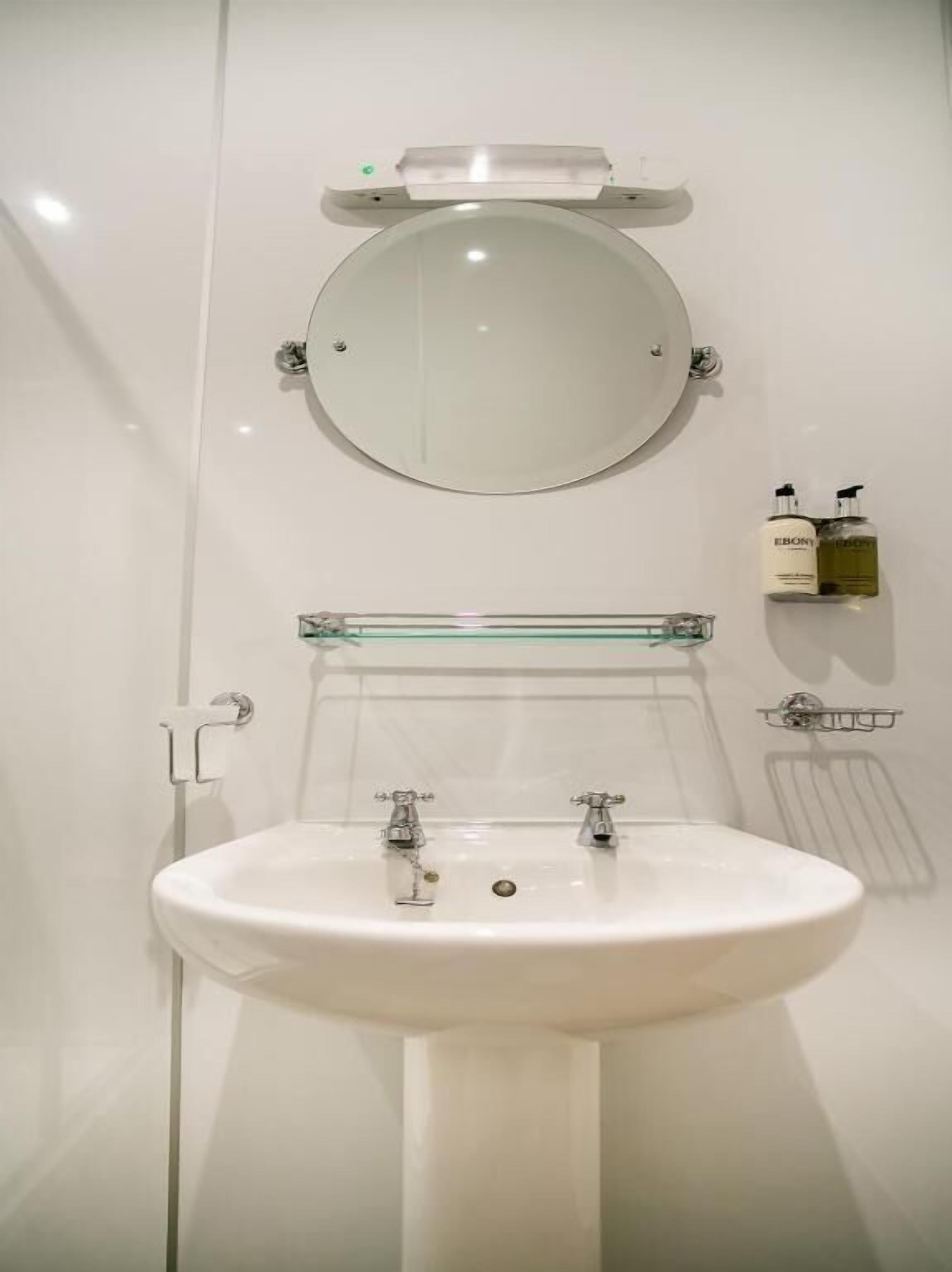 A bathroom with a white sink, a round mirror, and two faucets. There are two bottles on a shelf above the mirror.
