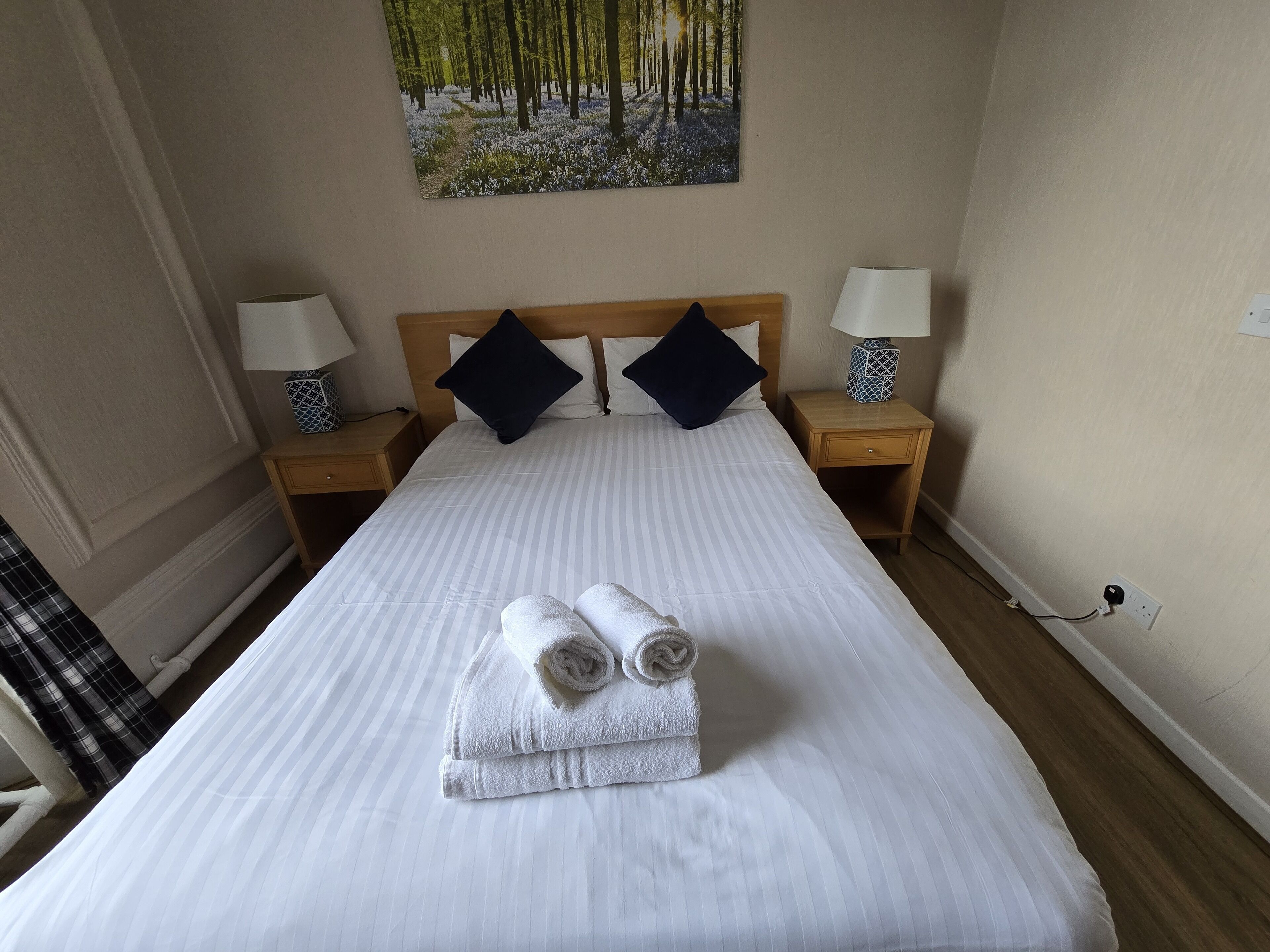 Una cama bien hecha con sábanas blancas y dos toallas enrolladas, flanqueada por dos mesitas de noche con lámparas, en una habitación con un cuadro de un bosque en la pared.