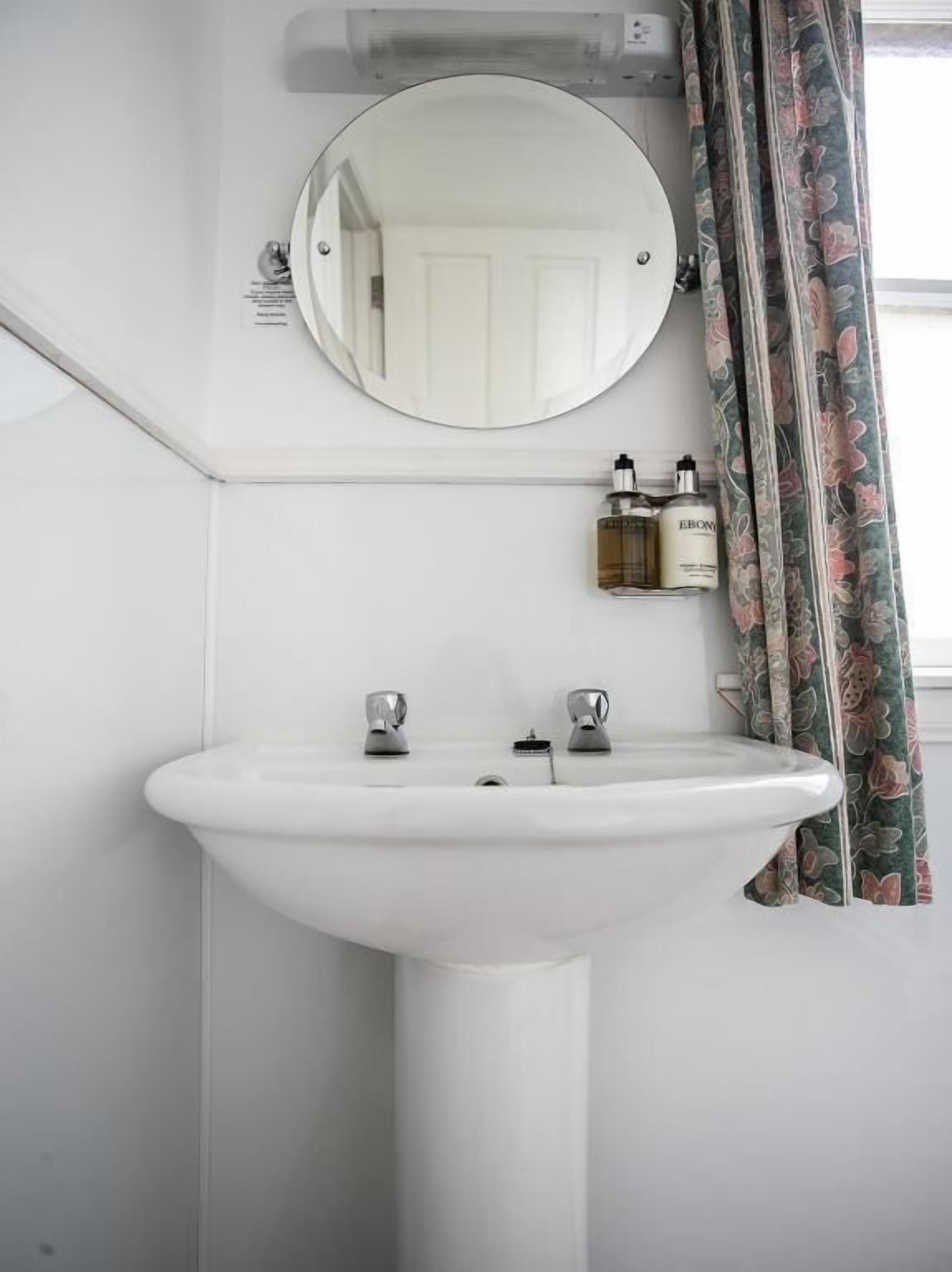 A bathroom with a white pedestal sink, a round mirror, and two bottles of toiletries on a shelf.