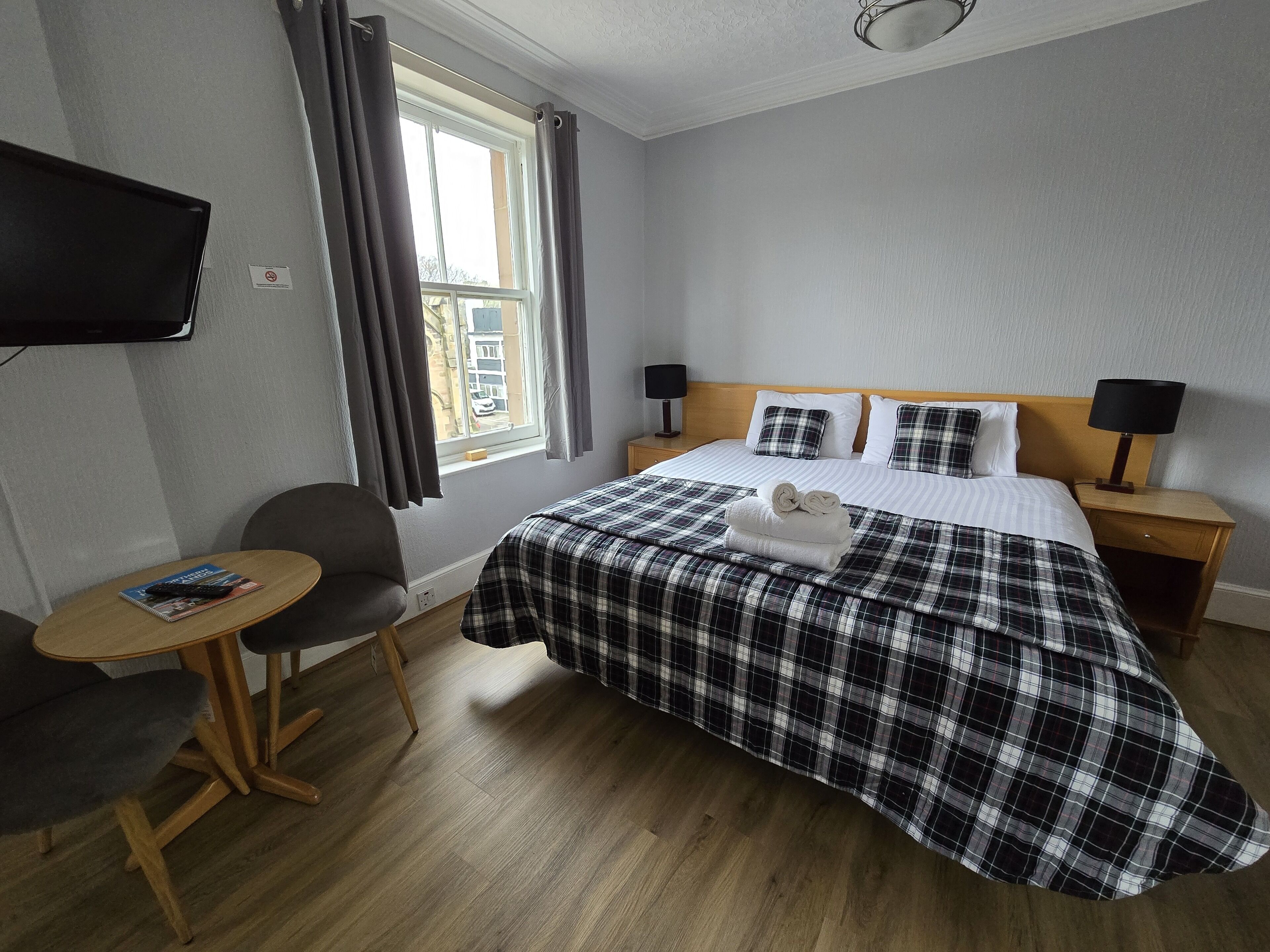 A hotel room with a bed, a TV mounted on the wall, a small table with a chair, and a window with curtains.