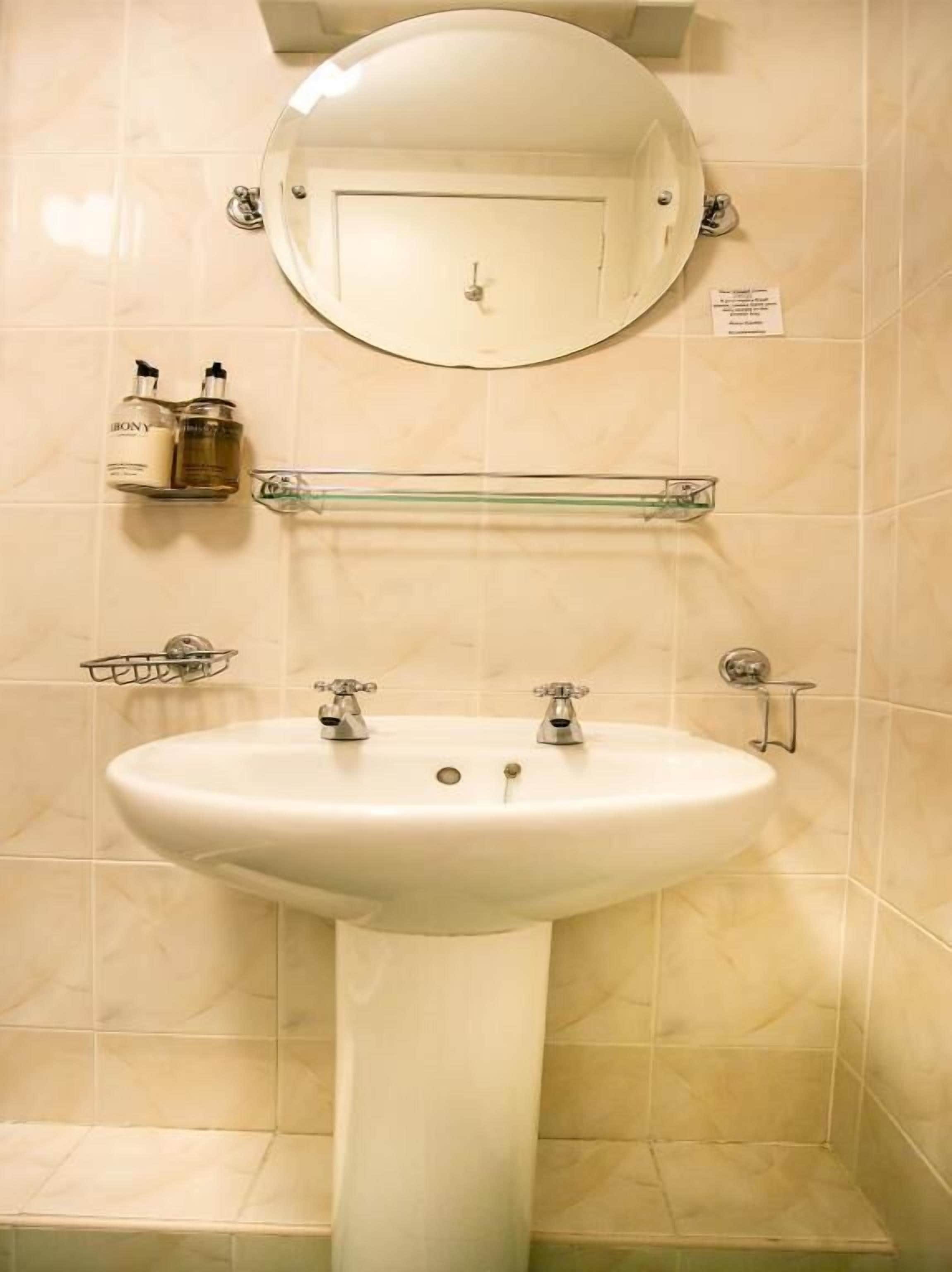 A bathroom with a pedestal sink, a round mirror, and two faucets. There are two shelves above the sink, one holding a bottle and the other a glass container.