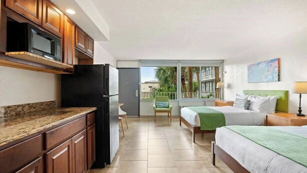 Quarto de hotel compacto com cozinha compacta, uma cama com colcha verde, mesa de cabeceira com luminária e vista para o exterior por uma porta de correr.
