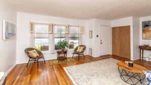 A room with hardwood flooring, two wicker chairs, a small table with a plant, and a view of the outdoors through a window with shutters. - Bay-View Bungalow, Dogs Welcome, Parking — Sleeps 9, Single Level (Ventnor City)