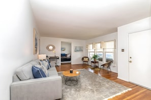 A modern living room with a sofa, coffee table, and a view through a window. - Bay-View Bungalow, Dogs Welcome, Parking — Sleeps 9, Single Level (Ventnor City)