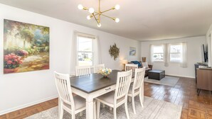 A dining area with a dark wooden table, white chairs, a chandelier, and a large painting of a garden scene. - Bay-View Bungalow, Dogs Welcome, Parking — Sleeps 9, Single Level (Ventnor City)