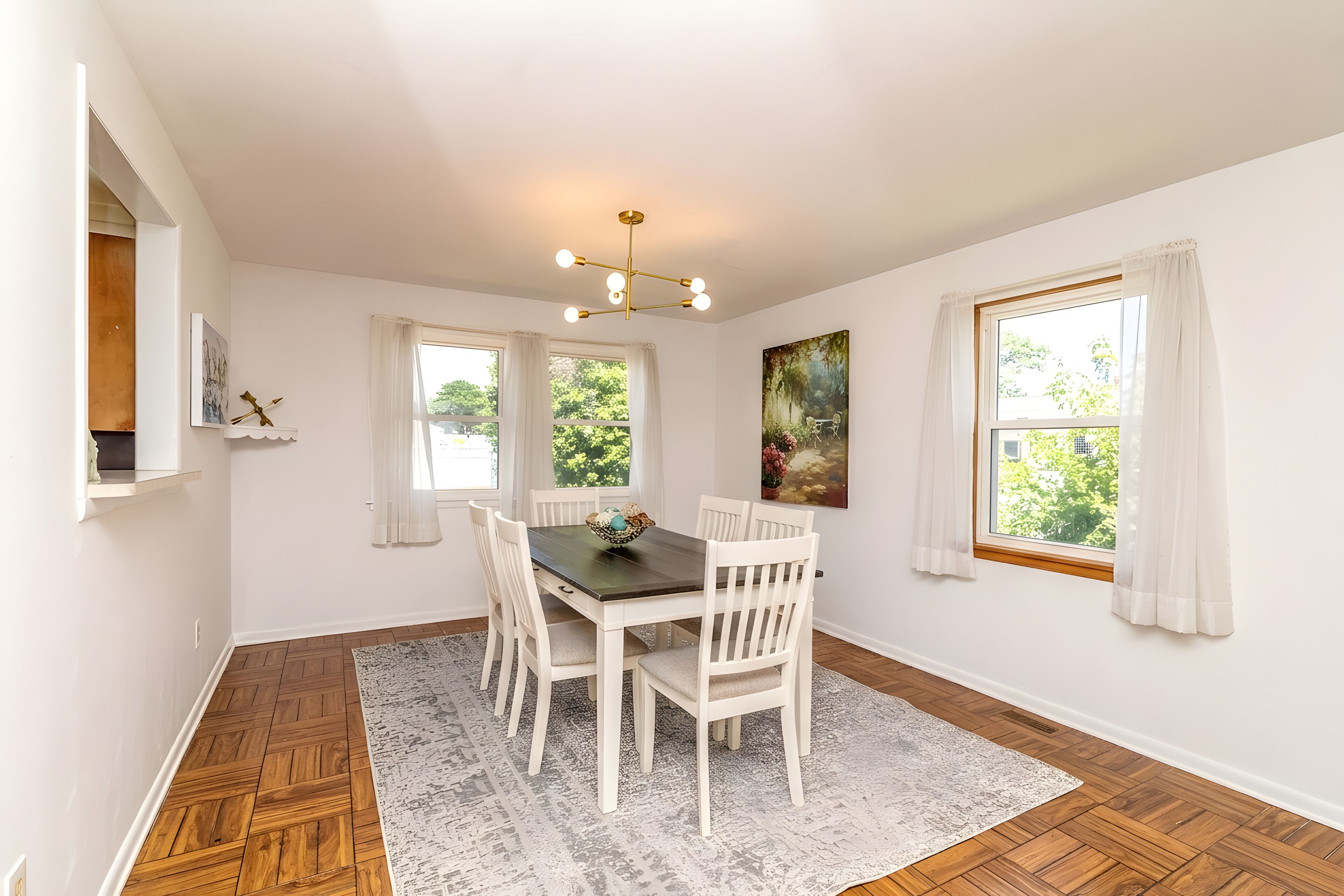 A dining area with a wooden floor, white walls, a dark wooden table, white chairs, a patterned rug, and a ceiling light fixture.