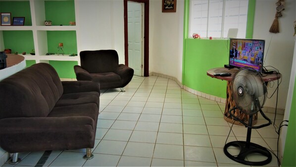 Una sala de estar con dos sofás oscuros, una pared verde, piso de baldosas, una puerta y un televisor sobre un mueble con un ventilador.