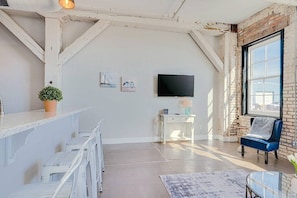 Une pièce spacieuse avec un plafond haut, des murs en briques apparentes, une grande fenêtre, un téléviseur à écran plat, un petit bureau et une chaise bleue.