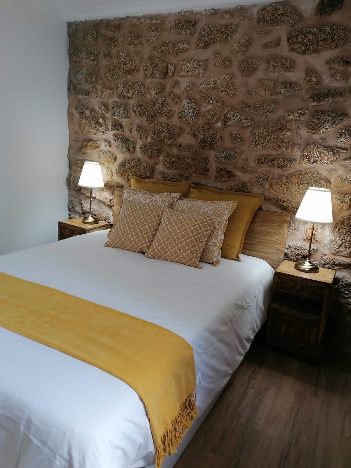 A bedroom with a stone wall, a bed with a white and yellow bedspread, two patterned pillows, and two bedside lamps.