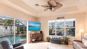 Ein geräumiges Wohnzimmer mit großem Fernseher, Deckenventilator, Blick auf den Pool und einer gemütlichen Sitzecke.