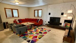 Un salon avec un canapé rouge, des fauteuils gris, une table basse, un tapis coloré, une cheminée et un téléviseur.