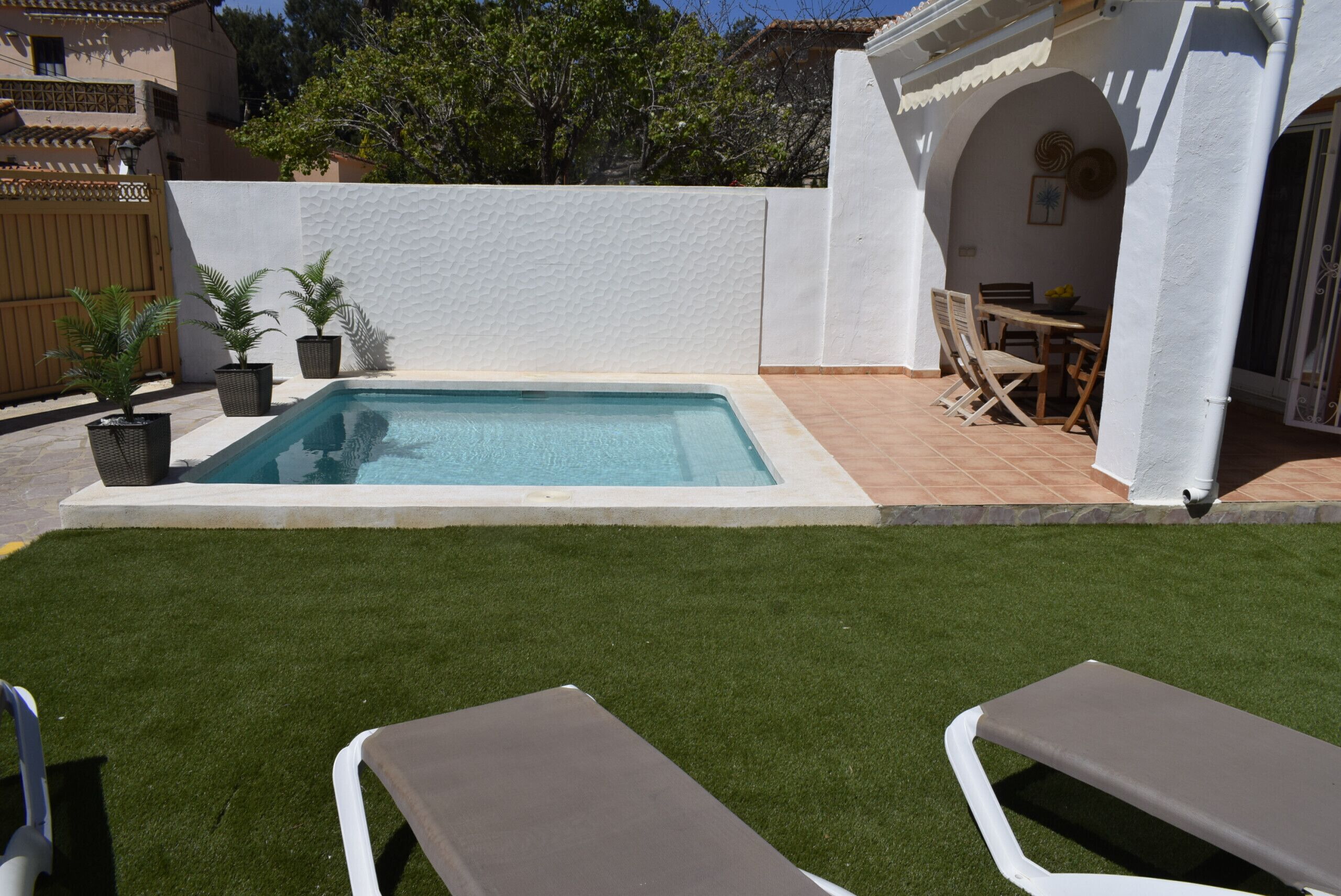 A swimming pool with a white wall and tiled floor, surrounded by artificial grass and two lounge chairs.