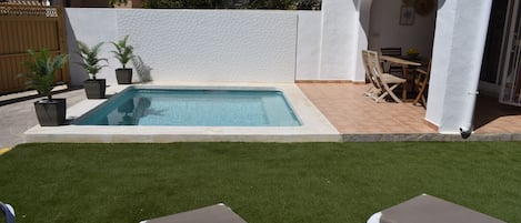 A swimming pool with a white wall and tiled floor, surrounded by artificial grass and two lounge chairs.