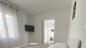 A bedroom with a bed, a bedside table with a lamp, a TV on the wall, and a window with curtains. - Apartments Roko - One bedroom apartment with balcony and sea view (A2) (Sveti Filip i Jakov)