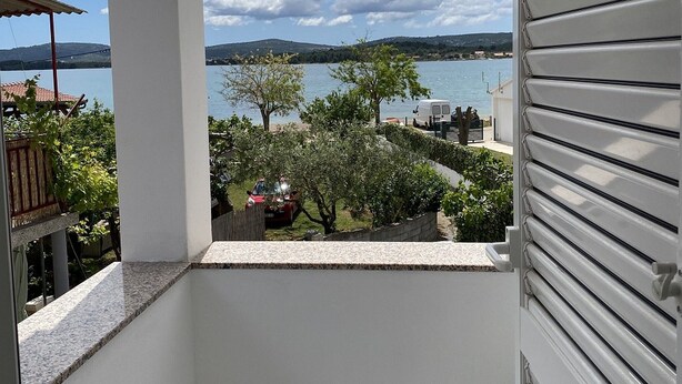 A balcony with a view of a lake and distant hills, a red car parked nearby, and a white door with horizontal slats. - Apartments Roko - One bedroom apartment with balcony and sea view (A2) (Sveti Filip i Jakov)