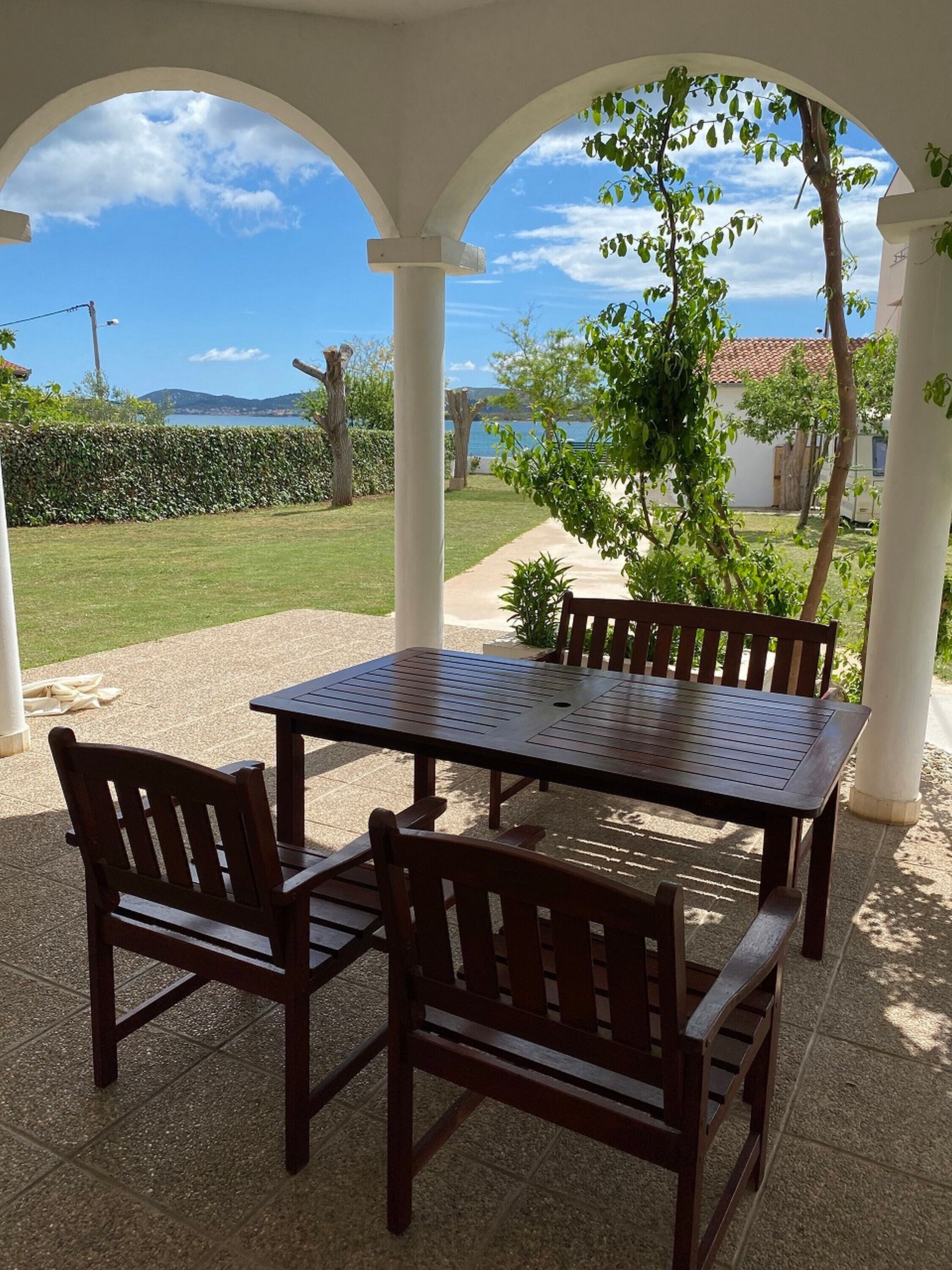 A covered outdoor patio with a table and chairs, a view of the sea, and a hedge in the background.