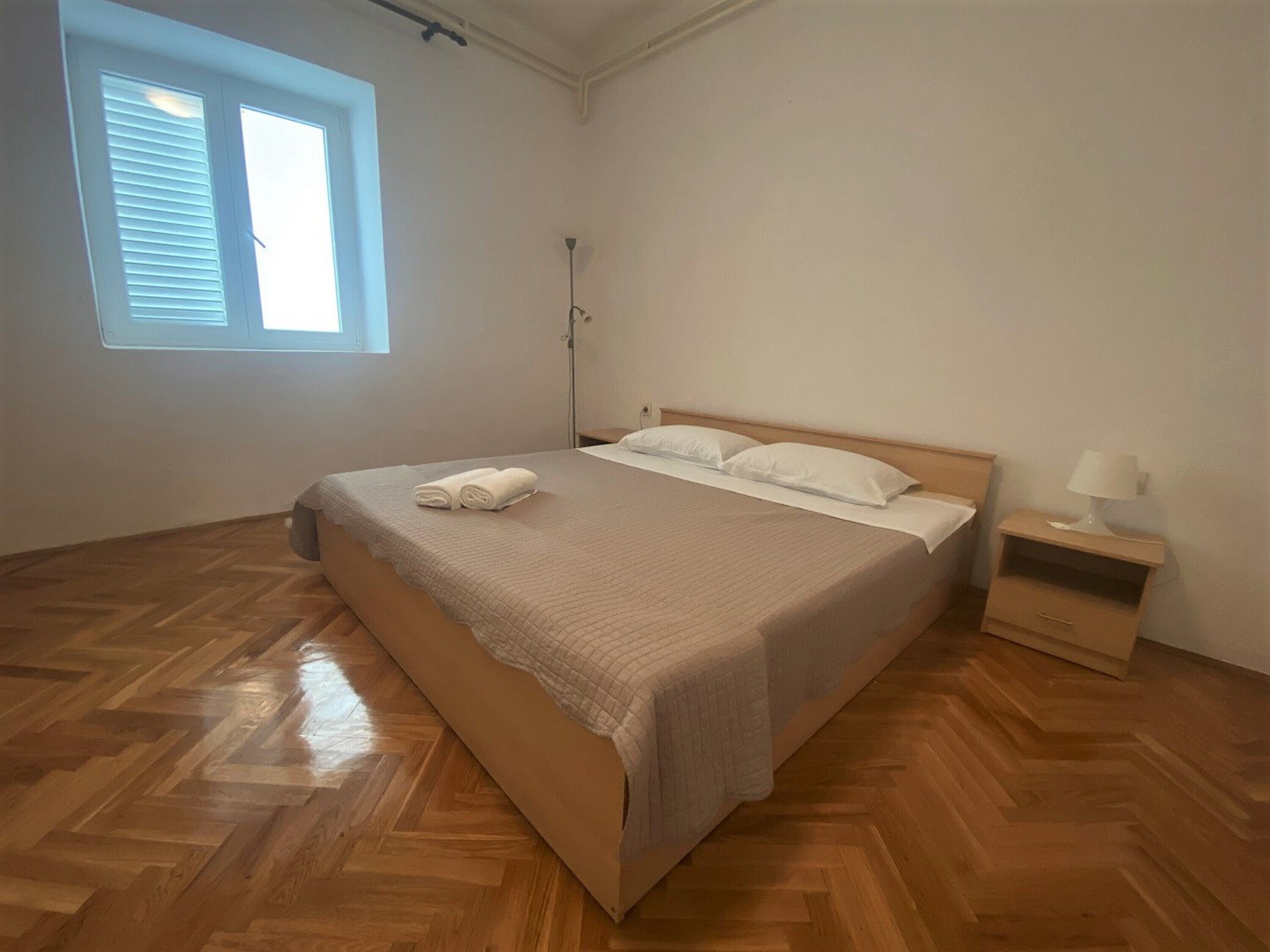 Un dormitorio con suelo de madera en espiga, una cama con sábanas blancas, una mesita de noche con lámpara y una ventana con persianas.
