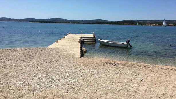 Ein Kiesstrand mit einer Betonsteganlage, die sich in ruhiges Wasser erstreckt, ein kleines Boot liegt am Ende festgemacht, und im Hintergrund sind sanfte Hügel unter blauem Himmel zu sehen.