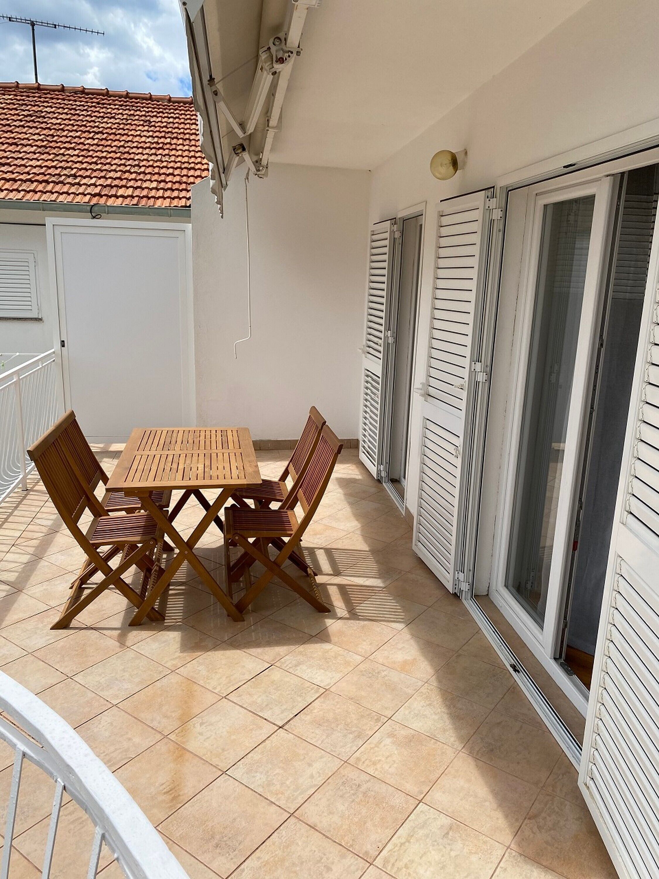 A balcony with a wooden table and chairs, white shutters, and a tiled floor.