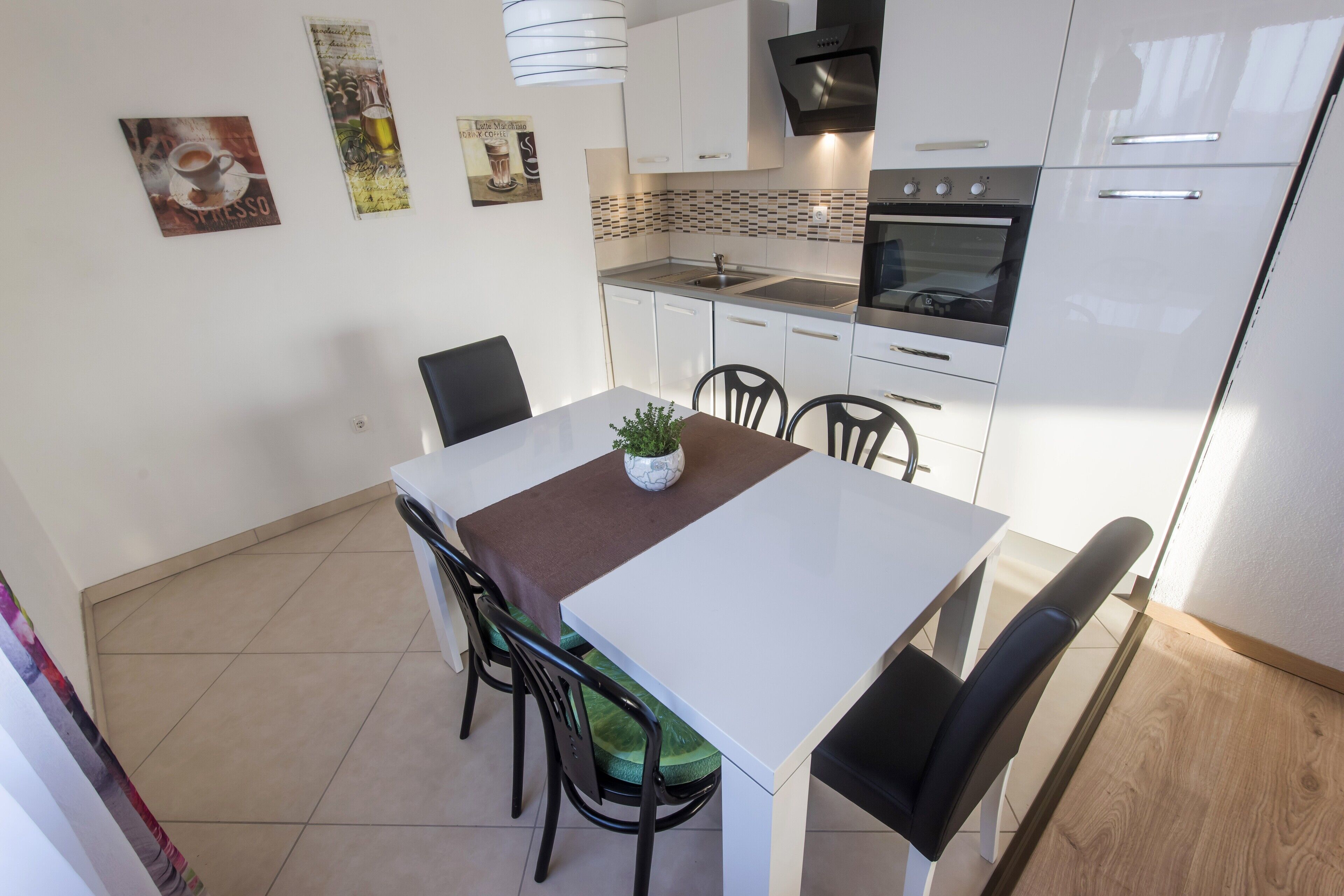 Une cuisine moderne avec des meubles blancs, un four encastré et un évier. On y trouve une table à manger avec des chaises et une petite plante en pot.