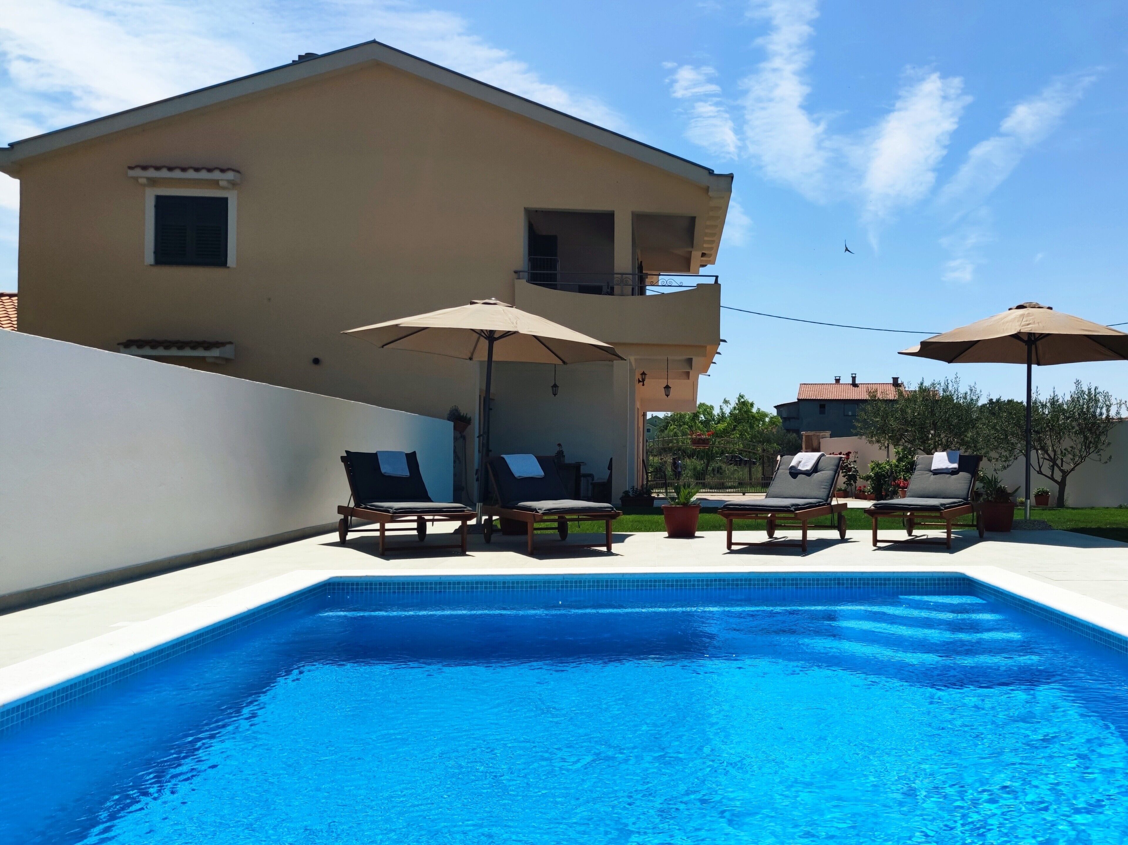Una piscina con sillas de descanso y sombrillas, un edificio color crema con balcón, y un cielo azul despejado.