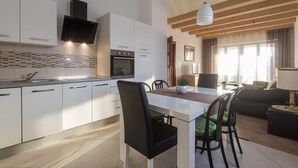 Una cucina moderna con mobili bianchi, un forno integrato e una cappa aspirante nera. È presente un'area pranzo con un tavolo bianco e sedie nere. La stanza ha travi a vista in legno e una lampada a sospensione.