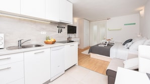 Cozinha moderna com armários brancos, lava-louças embutida e pia. Há um pequeno espaço de jantar com mesa e cadeiras. O quarto tem uma cama com roupa de cama branca e uma mesa de cabeceira com abajur.