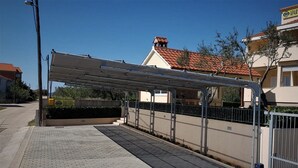 A covered parking area with a paved walkway, metal railing, and a building with a red-tiled roof in the background. - San Pedro Two bedroom apartment with sea view balcony (A2) (Sv. Petar na Moru)