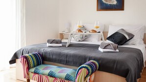 A neatly made bed with a gray bedspread, two pillows, and a bedside table with a lamp and a tray with croissants and orange juice.