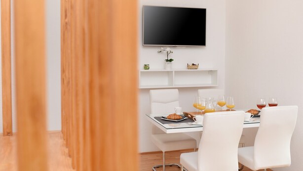 A modern dining area with a flat-screen TV mounted on the wall, white chairs, a glass table, and breakfast items including croissants and orange juice.