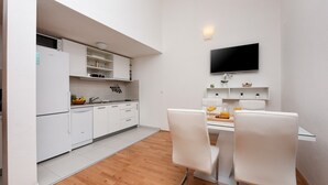 A compact kitchen with white cabinets, a dining area with two white chairs, and a flat-screen TV mounted on the wall.