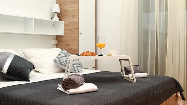 Een net opgemaakt bed met een donkere spreideken, kussens en een opgerolde handdoek. Naast het bed staat een klein tafeltje met een glas sinaasappelsap en een croissant.