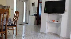 Une pièce comprenant un lit, une chaise en bois, un téléviseur, une petite étagère avec une télécommande et un objet décoratif, ainsi qu’une porte donnant sur une autre pièce.
