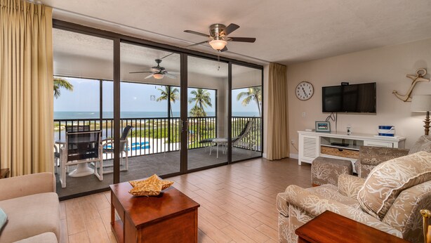 A spacious living room with a large sliding glass door leading to a balcony with a view of the ocean. There is a ceiling fan, a flat-screen TV, and a small white cabinet. - Now Open, Fully remodelled and restored (Fort Myers Beach)