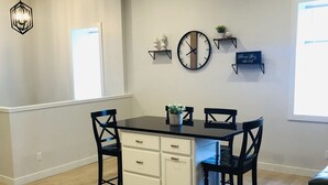A dining area with a black table and white cabinets, four chairs, a wall clock, and two shelves with decorative items. - Is This Heaven Downtown Dyersville Apartment (Dyersville)
