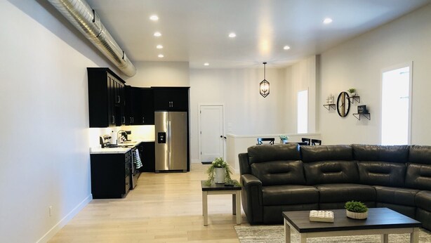 A modern living room with a black leather sofa, a coffee table, and a rug. There is a kitchen area with black cabinets and a stainless steel refrigerator. - Is This Heaven Downtown Dyersville Apartment (Dyersville)
