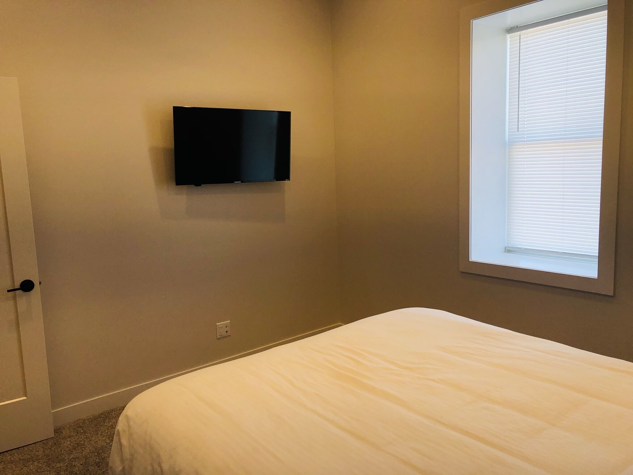 Une chambre à coucher comprenant un lit, un téléviseur fixé au mur, une fenêtre avec des stores et une porte.