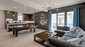 Ein modernes Wohnzimmer mit Billardtisch, einer Couch und einer Küchenzeile mit dunklen Schränken und einem großen Fenster.