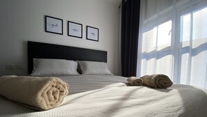 Una cama bien hecha con toallas enrolladas, un cabecero oscuro y tres palabras inspiradoras enmarcadas en la pared.
