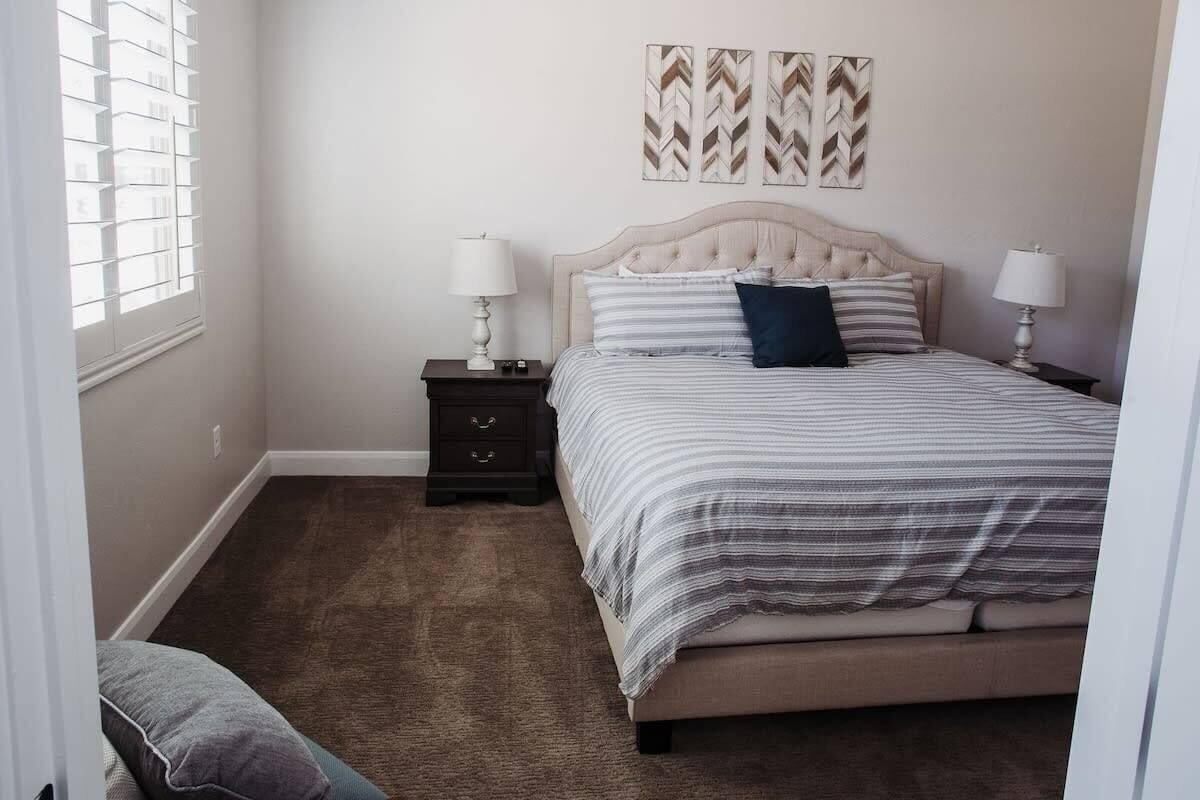 Un dormitorio con una cama, dos mesitas de noche, un mueble auxiliar, un ventanal con persianas y tres elementos decorativos colgados en la pared.