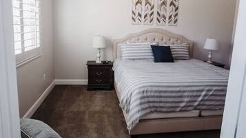 Un dormitorio con una cama, dos mesitas de noche, un mueble auxiliar, un ventanal con persianas y tres elementos decorativos colgados en la pared.